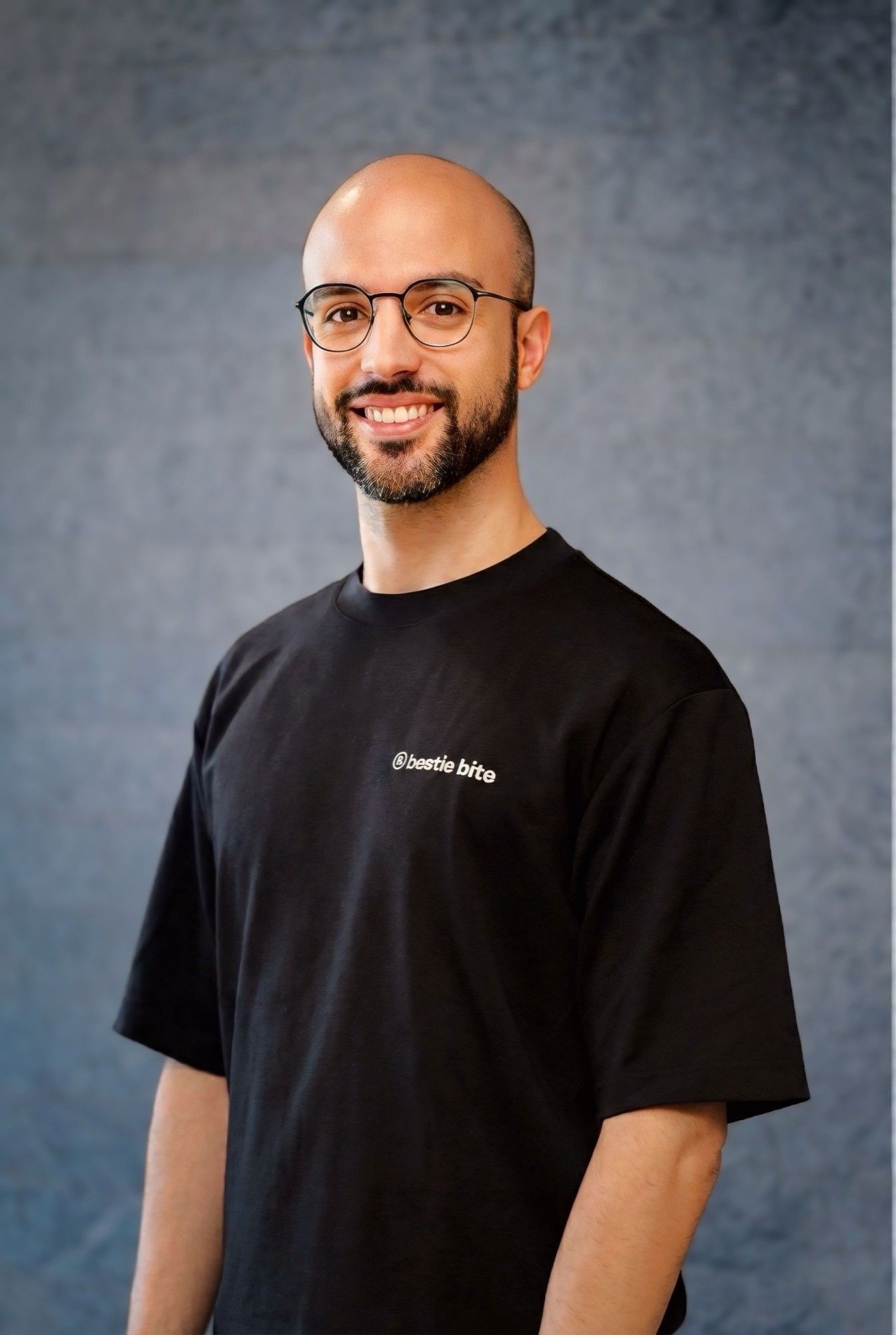 Portrait of Placido Falqueto: smiling, with a shaved head, beard and round glasses, wearing a black Bestie Bite T-shirt in front of a painted slate-blue studio backdrop.